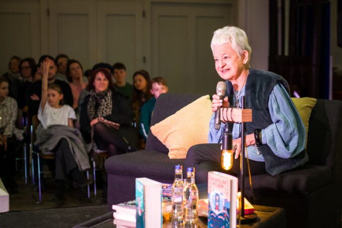 Female sitting on a grey sofa and talking to an audience.