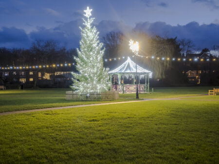 A park with a lit Christmas tree and a bandstand decorated with white lights
