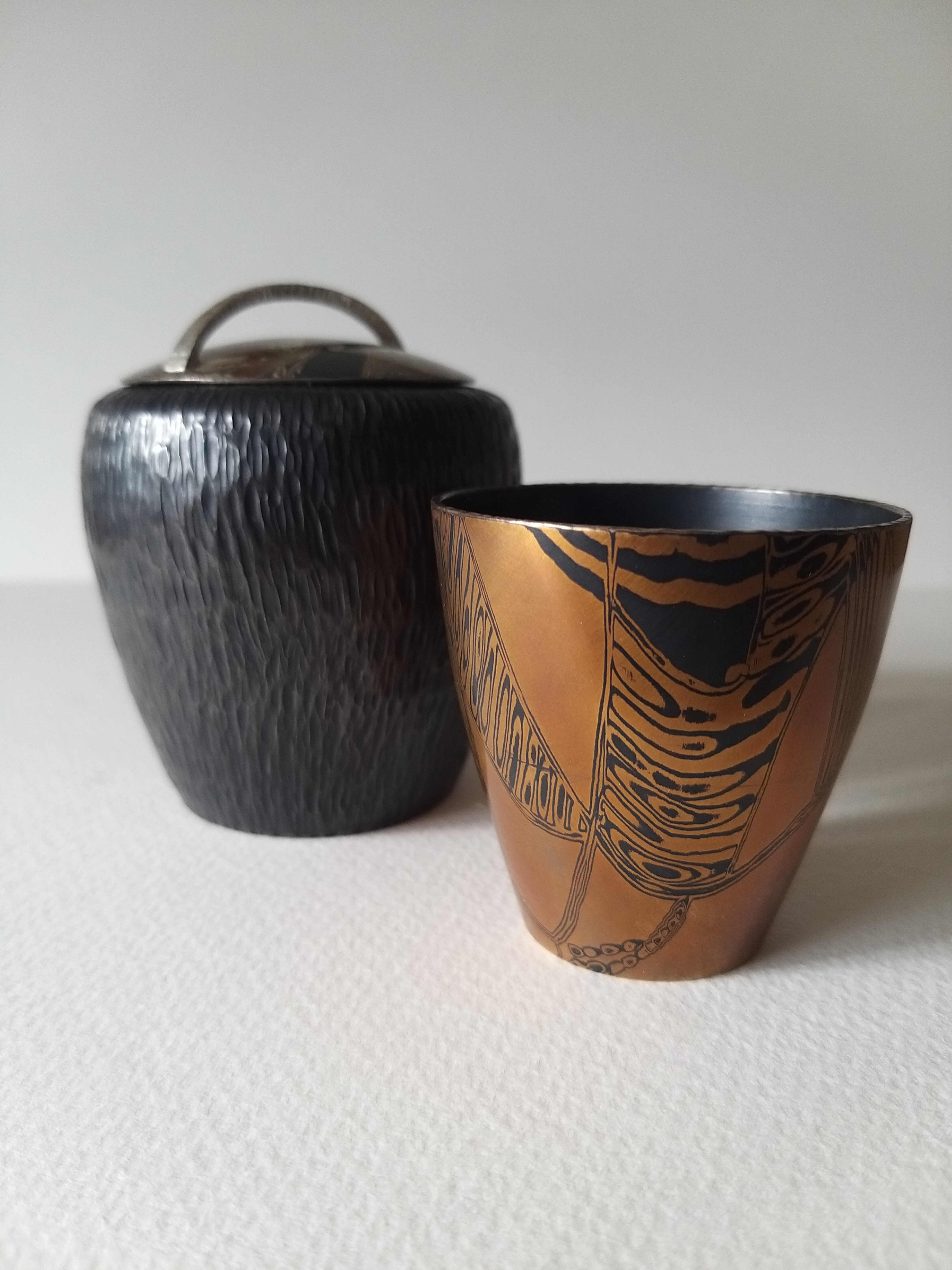 A small black vessel with a lid next to a bronze coloured cup.